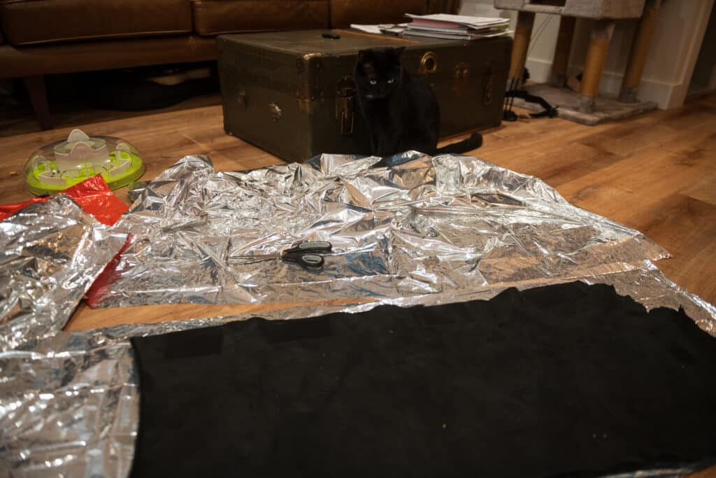 Cut out of SOL emergency blanket laying on the floor in a living room, prepared to be layered with sheepskin for insulating a cat backpack carrier.
