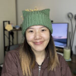 Close up of a woman wearing a cream colored crochet cat ear beanie, smiling slightly, with a blurred background