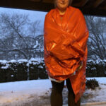 Lady standing outside in the cold winter weather. She is wrapped in emergency blanket, demonstrating its effectiveness against wind and cold for insulating a cat backpack carrier.
