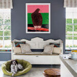 A brown and white spaniel on a green dog bed in a modern grey living room with bright colorful artwork, white painted shabby chic bench with assorted cushions, round wooden stool next to a countertop and large windows with view of garden behind, with beige and white striped blinds